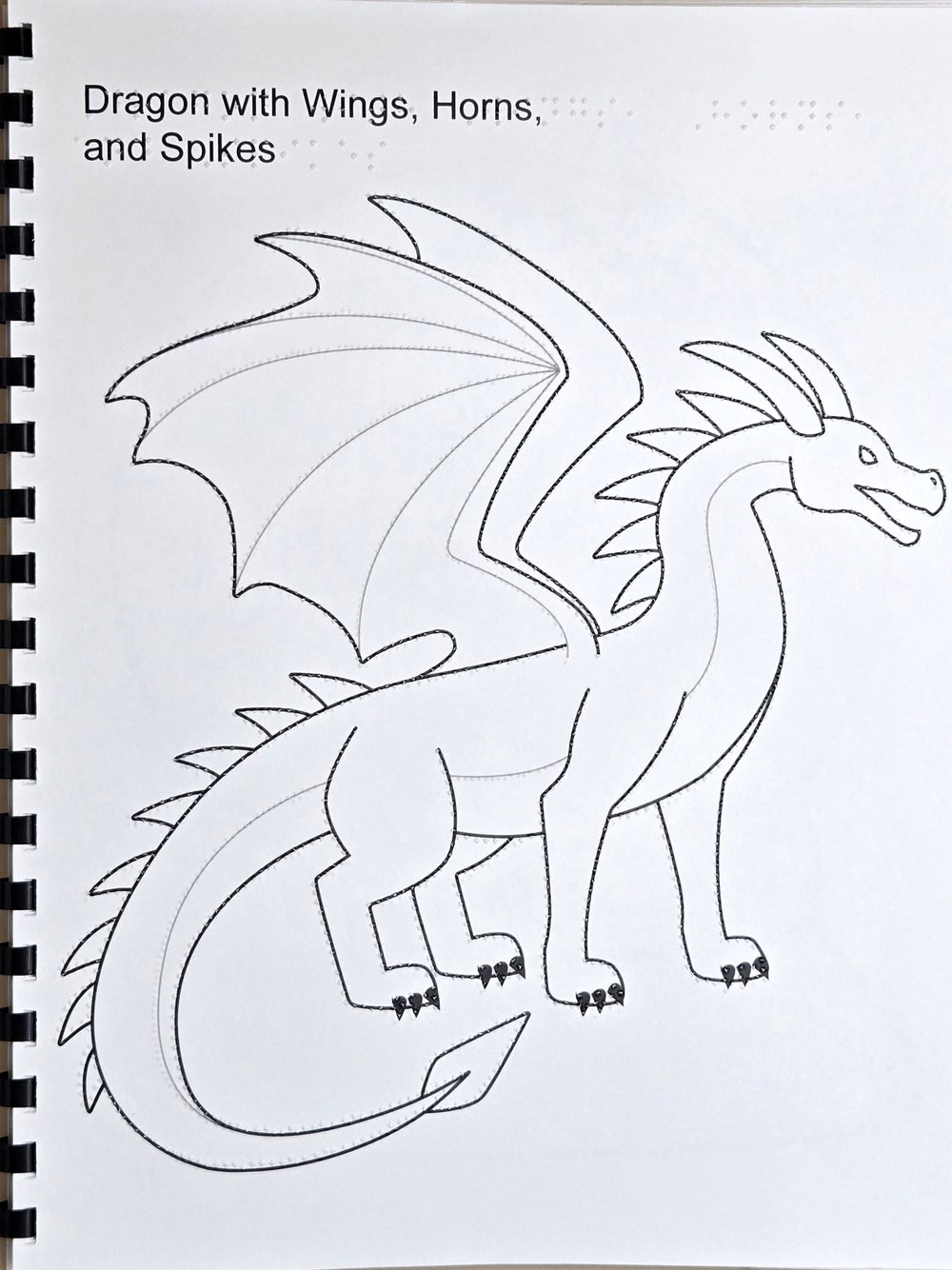 Page from a tactile coloring book: a dragon with wings, horns and spikes drawn with black and grey lines and raised dots along the lines
